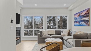 Ein modernes Wohnzimmer mit einem grauen Ecksofa, einem Kamin und großen Fenstern mit Blick auf schneebedeckte Bäume.