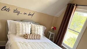 A neatly made bed with a quilted white comforter, a striped pillow, and several fluffy pillows. A small white stool with a vase of flowers is beside the bed. - Charming Waterfront Loft + NearShops+Scenic (Quinte West)