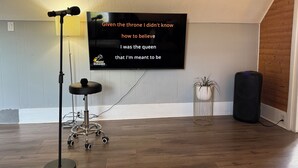 A karaoke room with a mounted TV displaying lyrics, a microphone stand, a speaker, and a potted plant. - Charming Waterfront Loft + NearShops+Scenic (Quinte West)