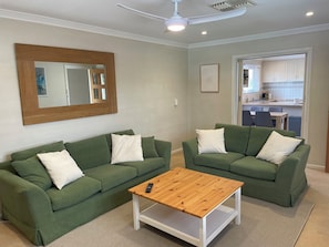 A living room with two green sofas, a wooden coffee table, a large mirror, and a kitchen visible through an open door. - Private and Secure, 3 bed 1.5 bath  (Kardinya)