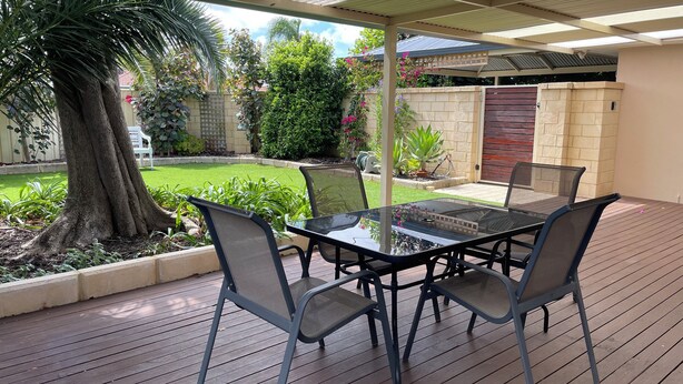 A covered patio area with a table and chairs, surrounded by a wooden deck and a garden with various plants and a tree. - Private and Secure, 3 bed 1.5 bath  (Kardinya)