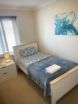A bedroom with a single bed, a bedside table with a lamp, a wall-mounted abstract painting, and a window with blinds. - Private and Secure, 3 bed 1.5 bath (Kardinya)