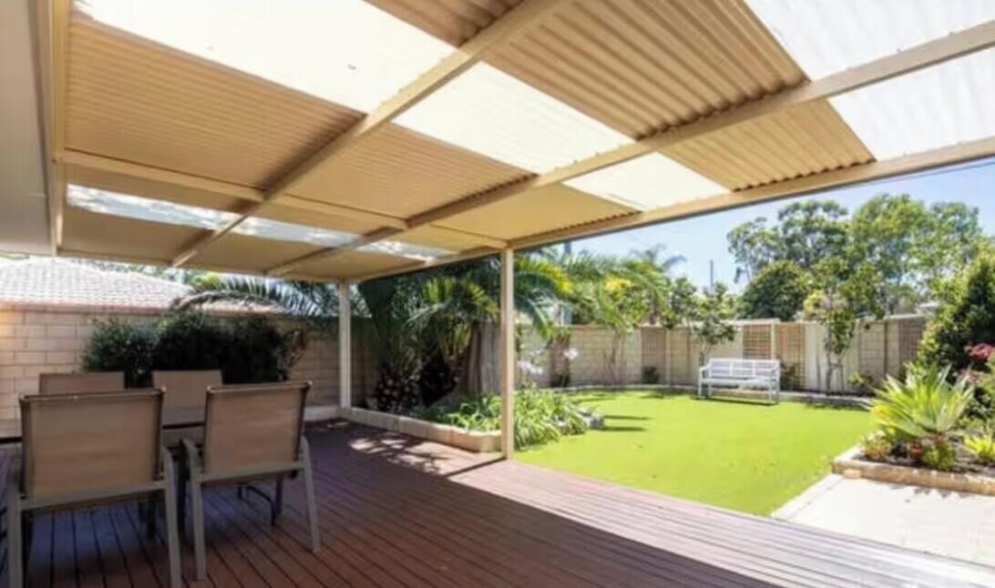 Uma área de varanda coberta com deck de madeira, mobiliário para área externa e vista para um gramado e jardim bem cuidados.