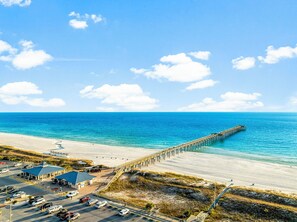 Une plage avec une jetée qui s’avance dans l’océan, un parking et un bâtiment avec un toit bleu.