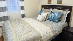 Una cama bien hecha con una colcha estampada y varias almohadas, una mesita de noche de madera oscura con cajones, y una ventana con cortinas de gasa.
