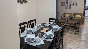 A dining area with a dark wooden table set for four, surrounded by chairs. There is a sofa in the background with cushions.
