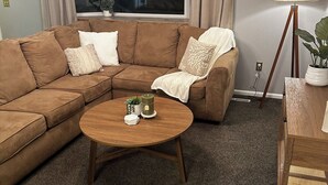 A living room with a brown sectional sofa, a round wooden coffee table, a ceiling fan, a window with curtains, and a floor lamp. - Charming 4-bedroom home with WiFi and massage chair in fabulous Colorado Springs (Colorado Springs)