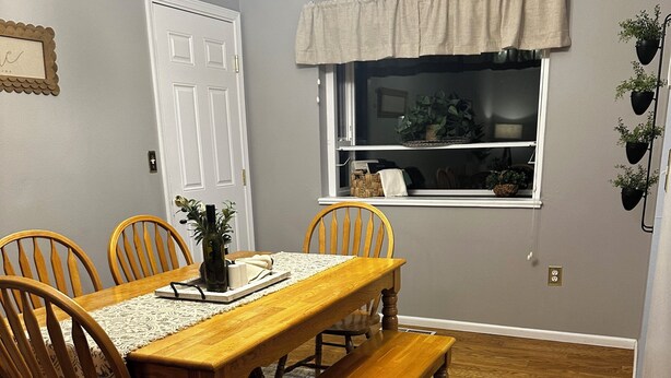 A dining room with a wooden table, four chairs, a bench, a rug, a window with a curtain, and a plant on the wall. - Charming 4-bedroom home with WiFi and massage chair in fabulous Colorado Springs (Colorado Springs)