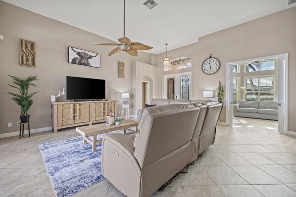 A spacious living room with a beige sofa, a flat-screen TV, a ceiling fan, and a large window with blinds.