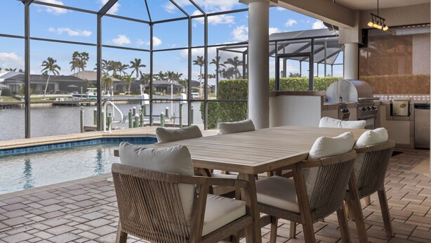 A poolside area with a wooden table and wicker chairs, overlooking a body of water with palm trees.
