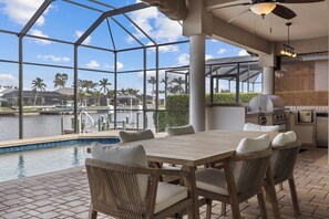 A poolside area with a wooden table and wicker chairs, overlooking a body of water with palm trees.