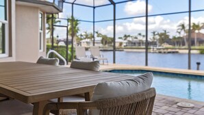A covered outdoor area with a pool, wooden table, wicker chairs, and a view of a body of water and palm trees.