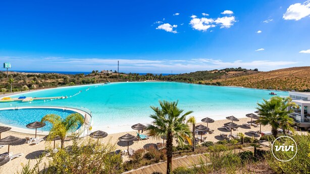 Un resort con una piscina grande, zona de playa con sombrillas y vistas al océano.