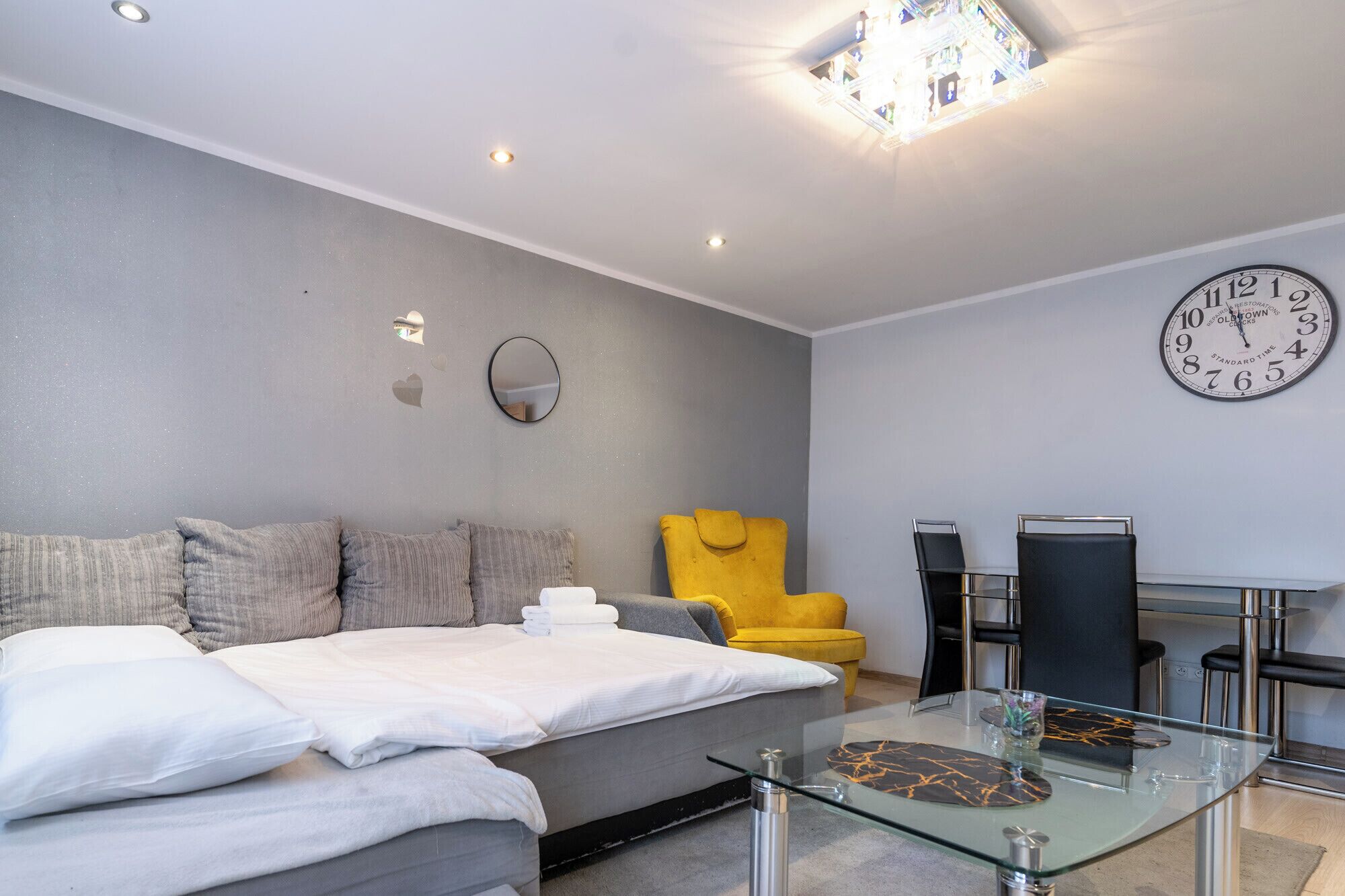 Una camera d'albergo moderna con un letto, una poltrona gialla, un tavolo da pranzo con sedie, uno specchio rotondo e una sveglia a muro.