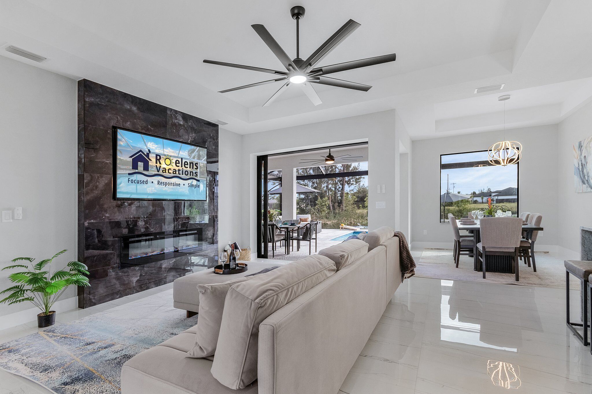 A modern living room with a large TV, a ceiling fan, and a dining area with a view of the outdoors.