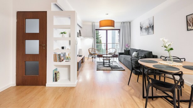 Ein modernes Wohnzimmer mit Holzboden, einer Couch, einem Esstisch mit Stühlen, einem Bücherregal und einem großen Fenster mit Vorhängen.