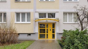 A building with a yellow entrance door, a brick pathway leading to it, and a small garden area with bushes and a tree.