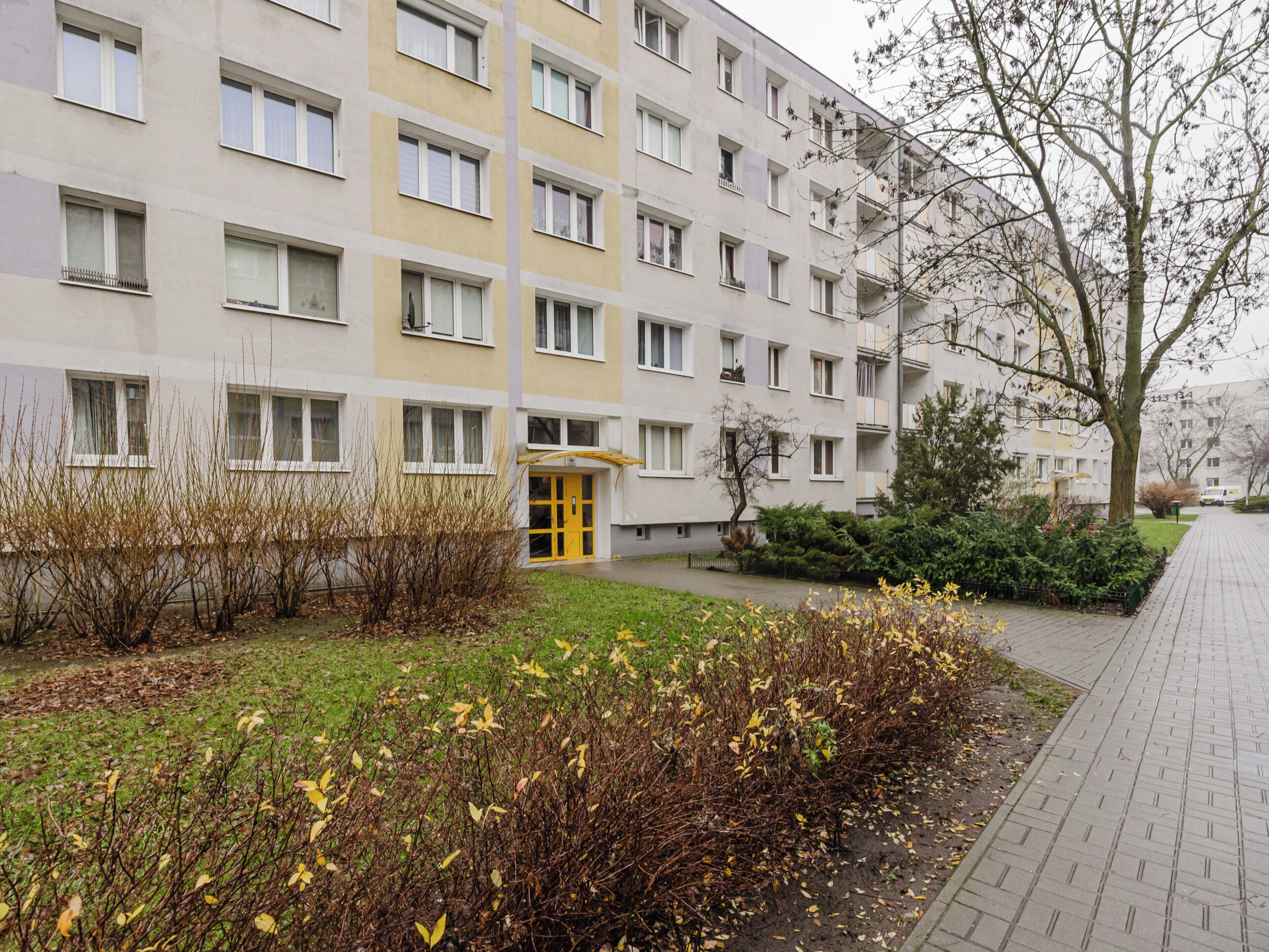 A multi-story residential building with a yellow entrance, surrounded by a garden with deciduous shrubs and a paved walkway.