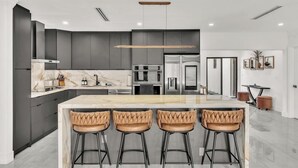 Una cucina moderna con mobili scuri, un piano di lavoro in marmo, elettrodomestici in acciaio inossidabile e tre sgabelli da bar in pelle.