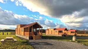 Eine Holzhütte mit Veranda und Stein Fundament, eingebettet in ein grünes Feld unter einem bewölkten Himmel.