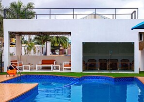 Uma área de piscina moderna ao ar livre com um edifício branco, espreguiçadeiras e uma área de estar coberta.