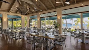 Un comedor amplio con varias mesas y sillas, ventanas grandes que ofrecen vista al mar, y un techo alto con vigas a la vista.