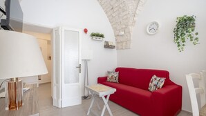 A red sofa, a white lamp, a clock, and a plant in a room with a white door and a stone ceiling. - GuestHost - Lovely apartment equipped with every comfort for 2 people and located in a typical stone building, of which it retains striking features in the internal architecture, such as the characteristic stone vault in the living area. The flat is loca (Ostuni)