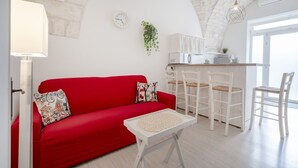 A red sofa, white chairs, a small table, a clock, and a plant in a room with a stone ceiling and a window. - GuestHost - Lovely apartment equipped with every comfort for 2 people and located in a typical stone building, of which it retains striking features in the internal architecture, such as the characteristic stone vault in the living area. The flat is loca (Ostuni)