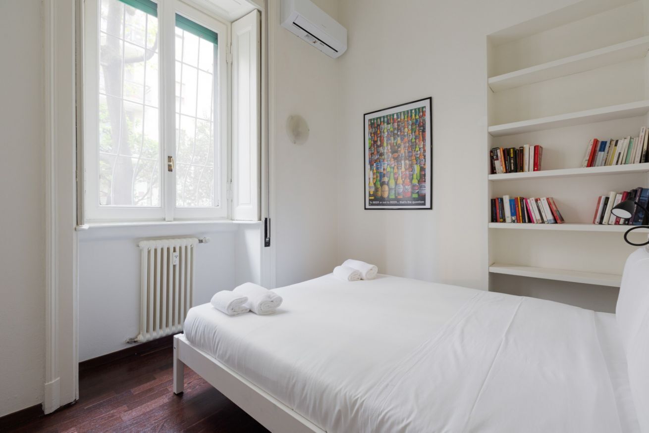 A bedroom with a bed, a radiator, a window with blinds, a bookshelf, and a framed picture on the wall.