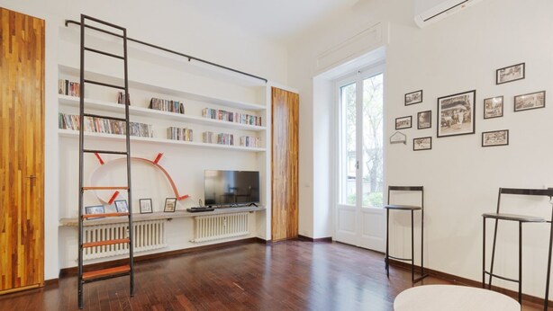 Una habitación moderna con piso de madera, una estantería, una escalera, un televisor y cuadros en la pared.