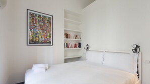 Una cama bien hecha con ropa blanca, una estantería con varios libros y un cartel enmarcado en la pared.