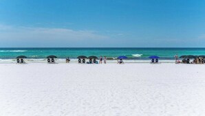 Una spiaggia con sabbia bianca, cielo azzurro e onde oceaniche. Si vedono ombrelloni e lettini.