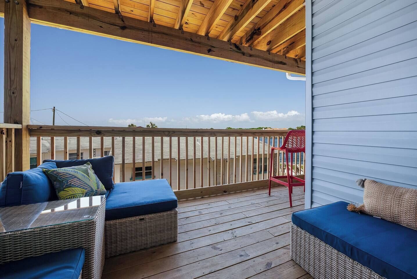 Une terrasse extérieure couverte, aménagée avec des meubles en osier et une chaise rouge, qui offre une vue sur une étendue d’eau.
