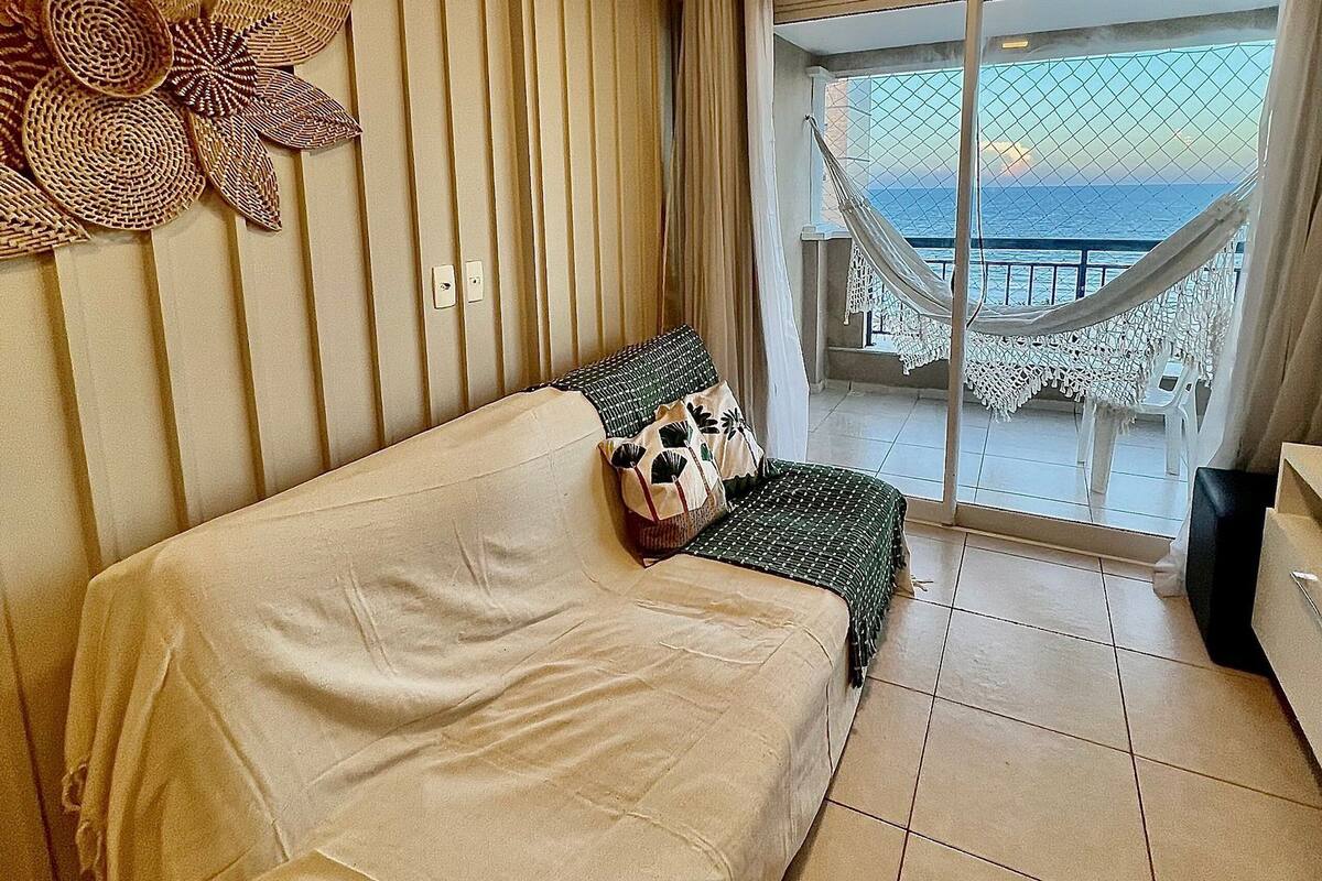 Une pièce avec un canapé beige, un panneau décoratif mural, une vue sur la mer par une porte vitrée et un balcon avec une chaise blanche.