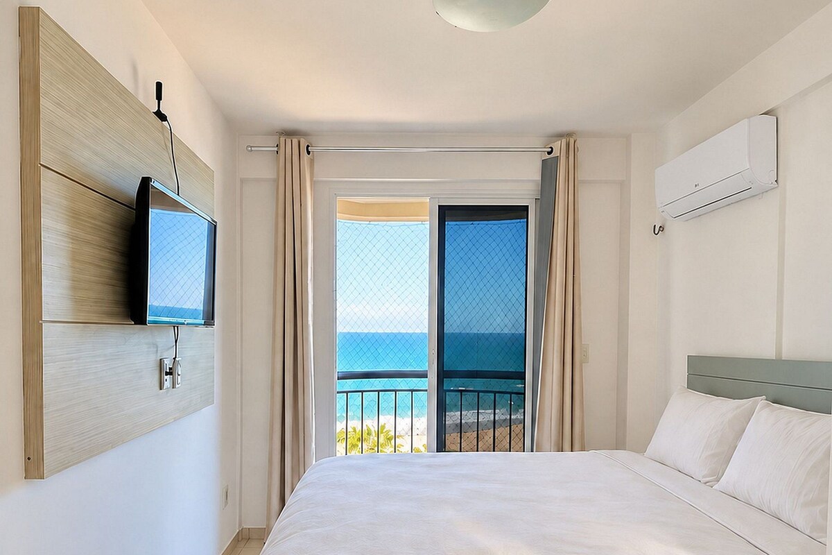 Una camera d'albergo con un letto, una televisione a muro, un condizionatore e vista sull'oceano da una porta finestra.