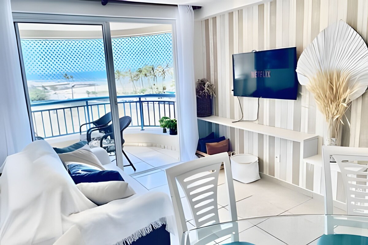 Un soggiorno moderno con un balcone vista spiaggia, una televisione a schermo piatto montata a parete e un tavolino da caffè in vetro.