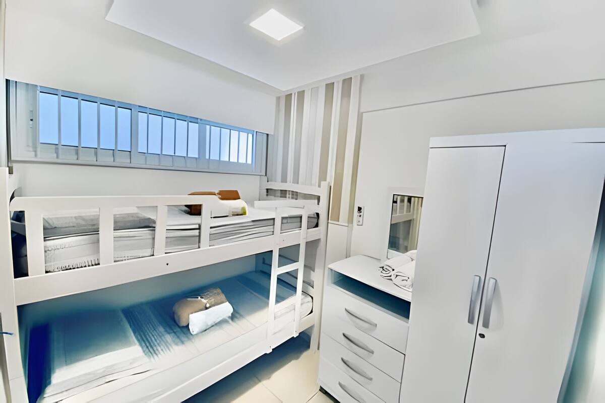Une chambre avec un lit superposé, une fenêtre, une armoire blanche et un bureau avec des tiroirs.