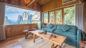 Ein Zimmer aus Holz mit Bergblick, ein grünes Sofa, ein Holztisch mit Teeservice und ein Fenster mit Vorhängen.
