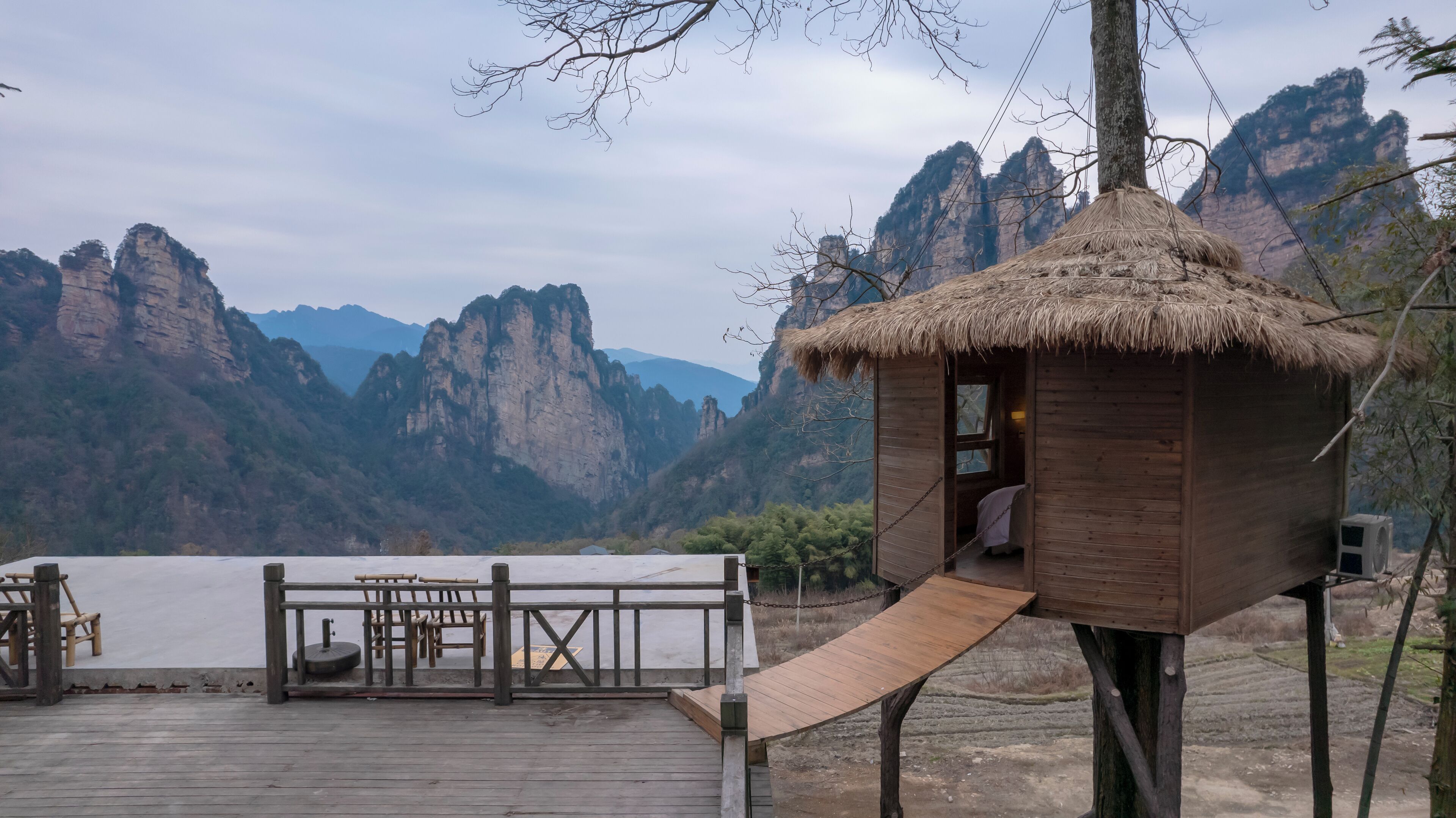 A wooden structure with a thatched roof, a wooden deck with chairs and a table, and a scenic view of mountains in the background.