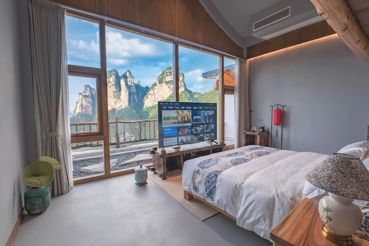 Un dormitorio con una ventana grande que ofrece vistas a las montañas, una cama con colcha estampada de motivos florales, una mesita de noche de madera con lámpara y un televisor en la pared.