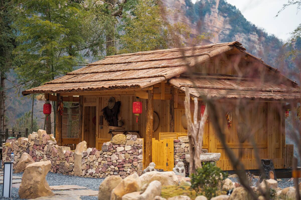 Una cabaña de madera rústica con techo de paja, rodeada por un muro de piedra y un jardín de rocas.