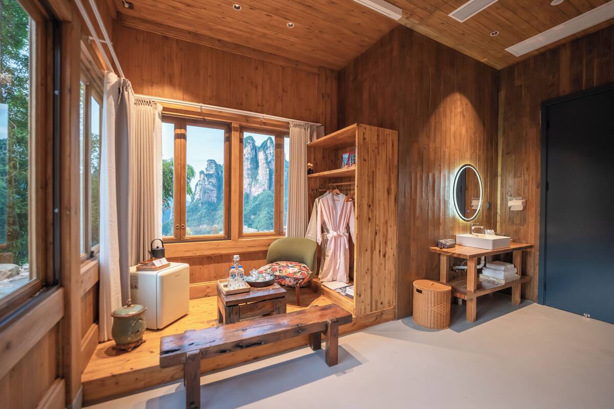 Interior de una cabaña de madera con un ventanal grande, un banco de madera, una mesa pequeña con una tetera, una silla, un espejo y un lavabo.