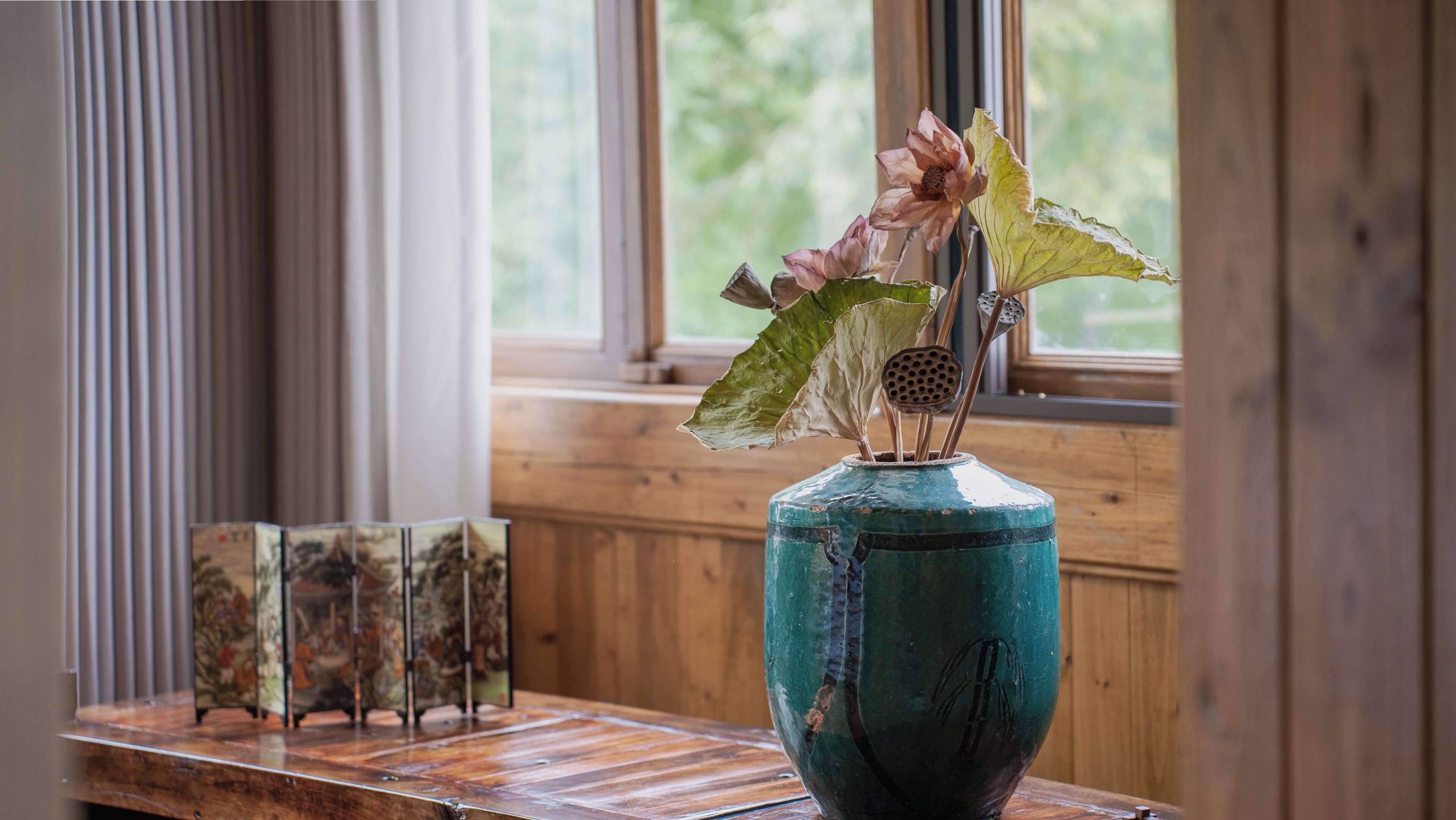 Un jarrón azul verdoso con flores secas y hojas sobre una mesa de madera cerca de una ventana con persianas.
