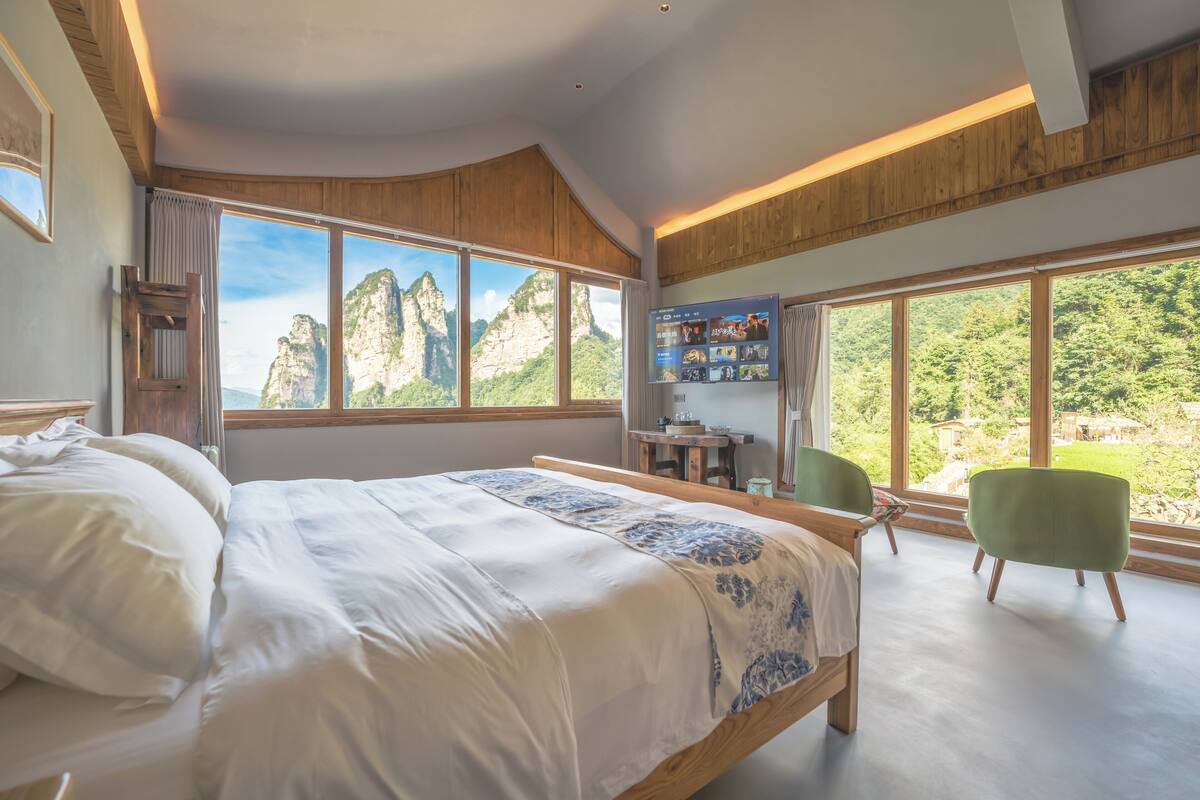 Habitación de hotel moderna con una cama grande, un escritorio y una silla. Amplias ventanas con vistas a montañas y follaje.