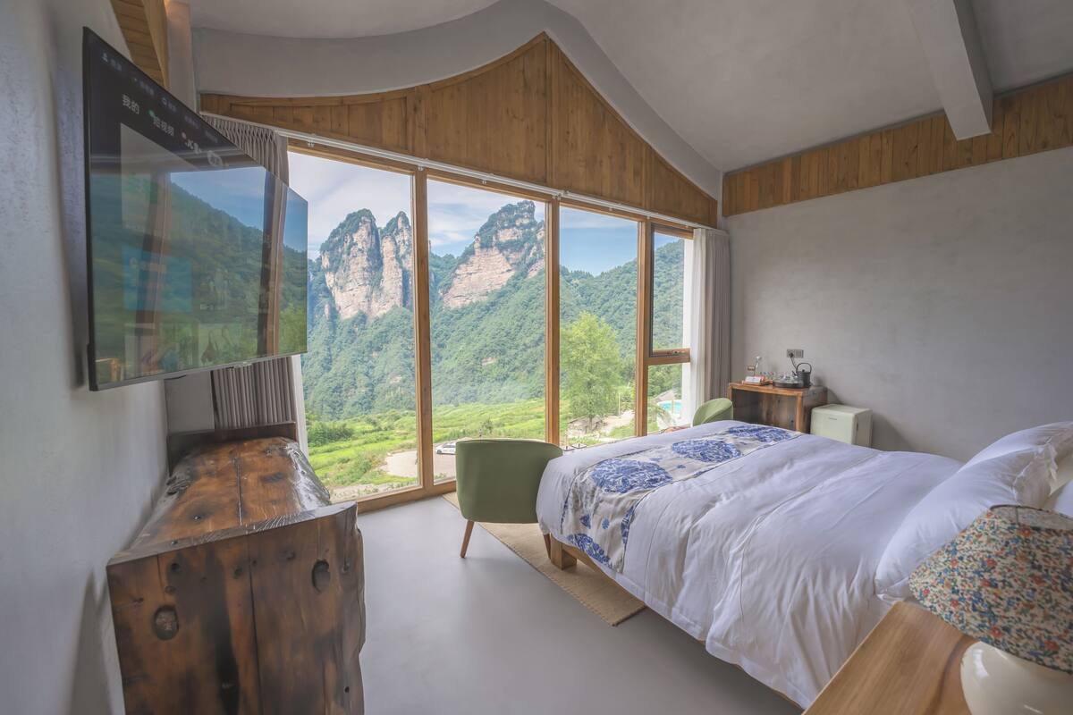 Un dormitorio con una ventana grande que ofrece vistas a la montaña, una cama, una silla y un aparador de madera.