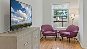 Una habitación con televisión en un mueble, dos sillas, una ventana con persianas y una lámpara de pie.