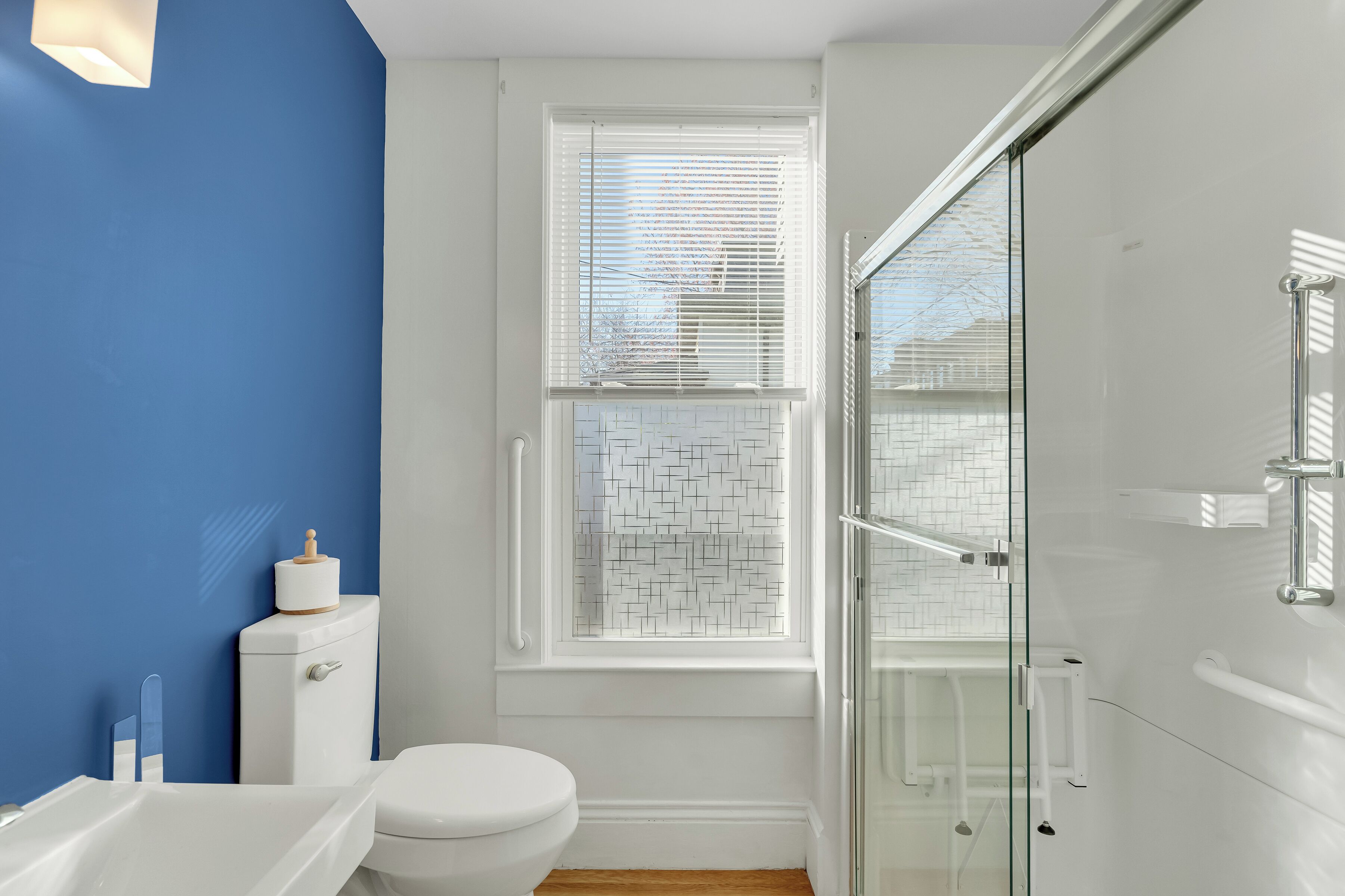Une salle de bain avec des toilettes blanches, une douche en verre et une fenêtre avec des stores blancs.