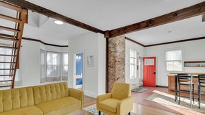 Un salon avec un canapé jaune, un fauteuil jaune et une table basse en bois. On y trouve un mur en briques, des poutres en bois au plafond et une porte rouge à l’arrière-plan.