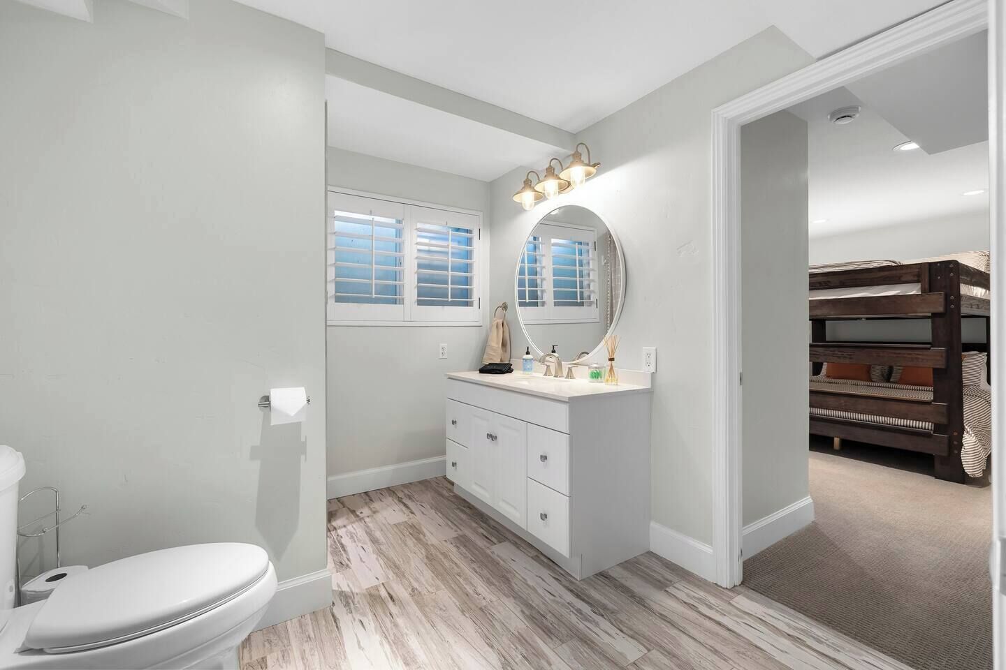 Een badkamer met toilet, wastafel met spiegel en een wastafelmeubel. Er is een raam met zonwering en een deur naar een andere kamer.