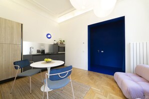 A modern living room with a round table, two chairs, a sofa, and a kitchen area with a blue door.